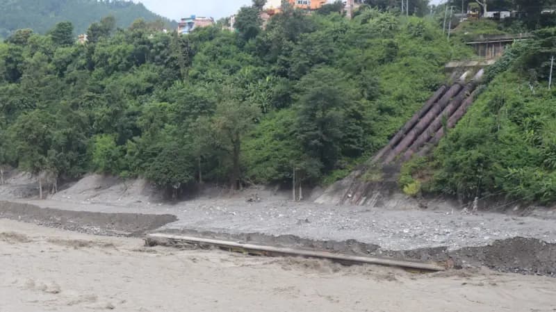 Swollen floodwaters inundate the powerhouse and penstock pipe area of a riverbank hydropower plant.