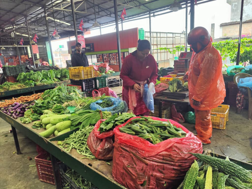 Fresh green vegetables including okra and gourds displayed at a market stall as a shopper in an orange raincoat looks on.