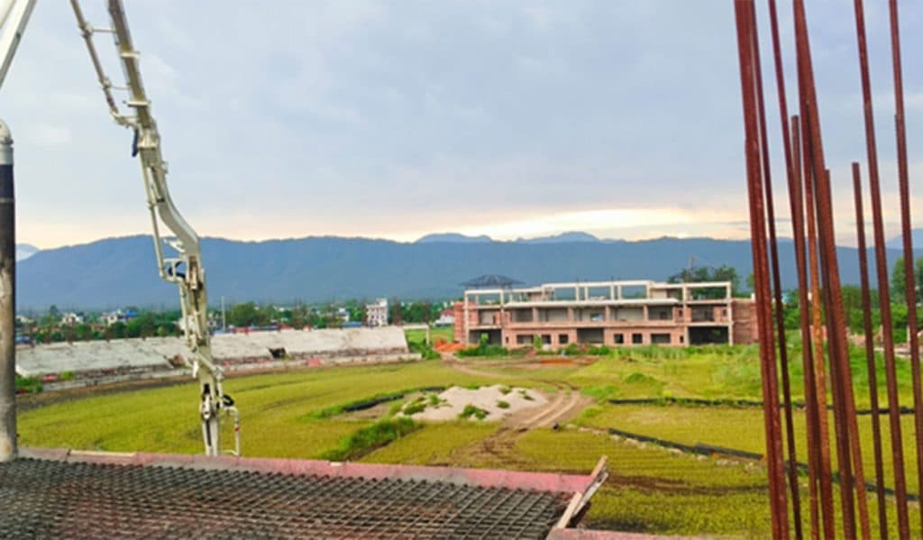 A wide view of the Gautam Buddha International Cricket Stadium construction site with unfinished structures and a concrete pump.