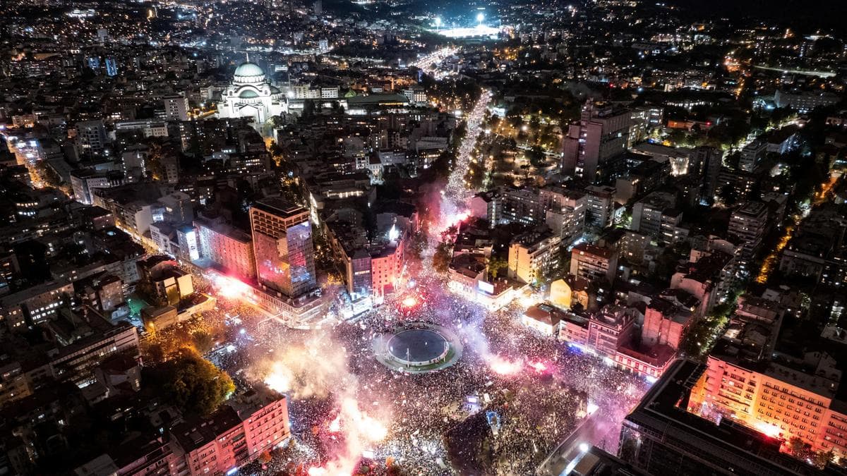 An aerial view of a large crowd in a city square at night, illuminated by flares.