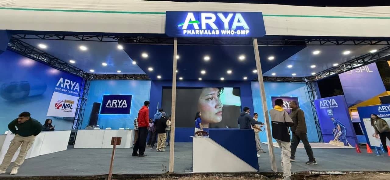A large, illuminated promotional booth for Arya Pharmalab at a stadium, with people walking in front of it.