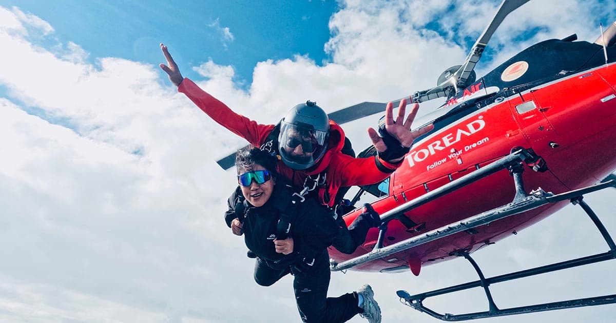 Two people in skydiving gear free-falling with a red helicopter in the background.