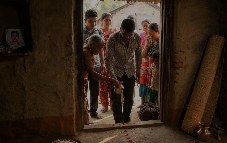 A family performs a ritual at their home doorway, with a framed portrait of a man visible on the wall.