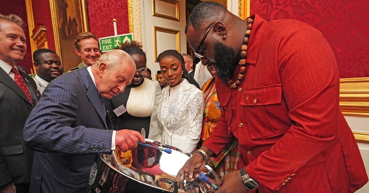 King Charles III interacting with a man playing a steel pan drum at an indoor event.