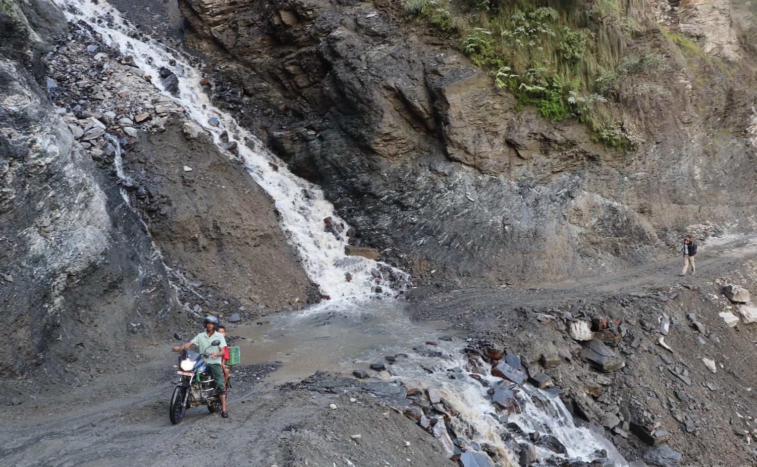 A motorcyclist navigates past a stream of water flowing across a damaged mountain dirt road.