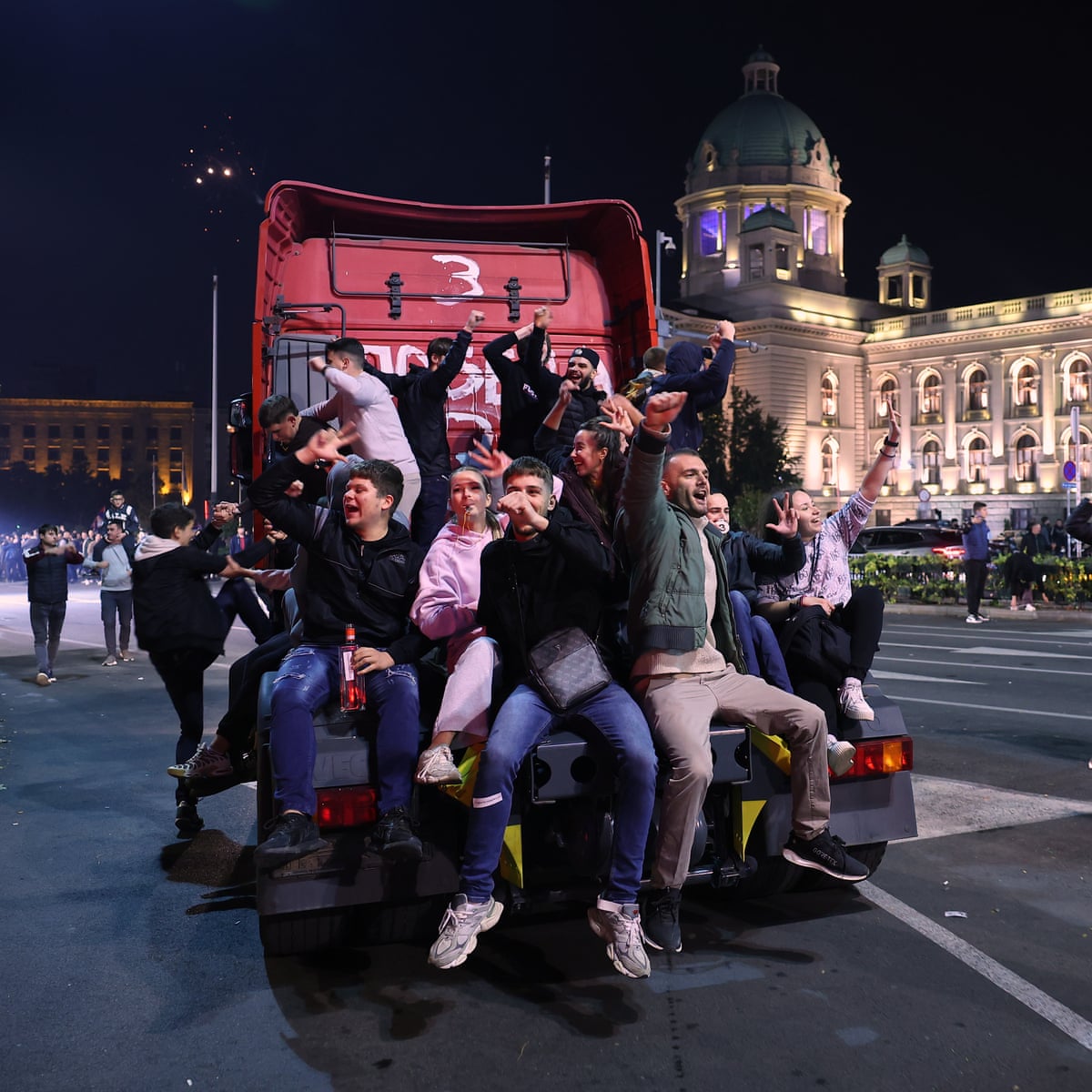 A group of young people celebrating on the back of a truck in front of a government building at night.
