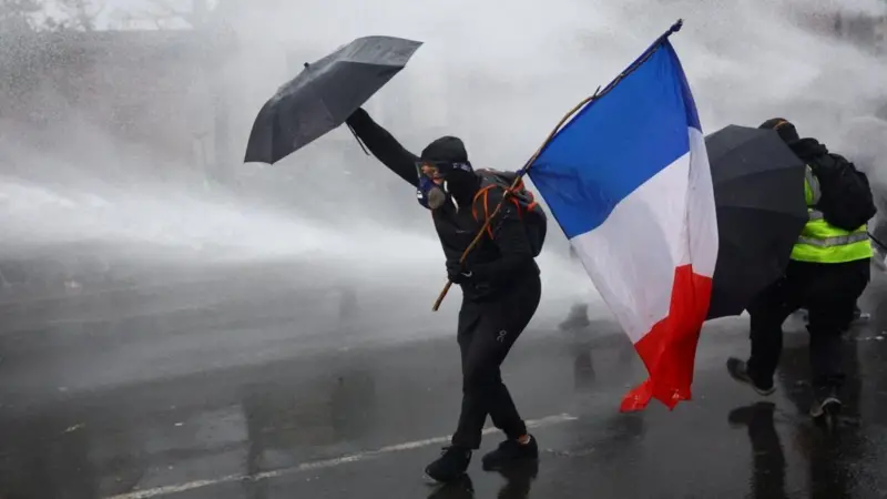 A protester holding a French flag and an umbrella while being sprayed by a water cannon.