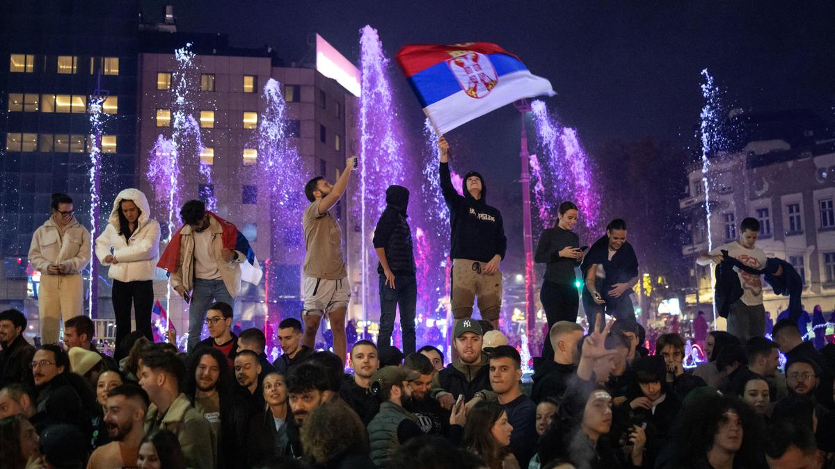 A group of young people celebrating at night with a Serbian flag in front of illuminated fountains.