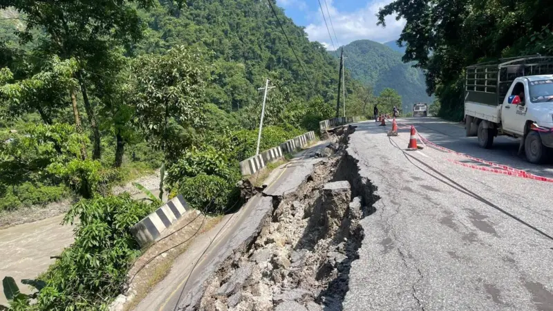 A collapsed edge of a paved highway cordoned off by safety cones and tape beside a river.