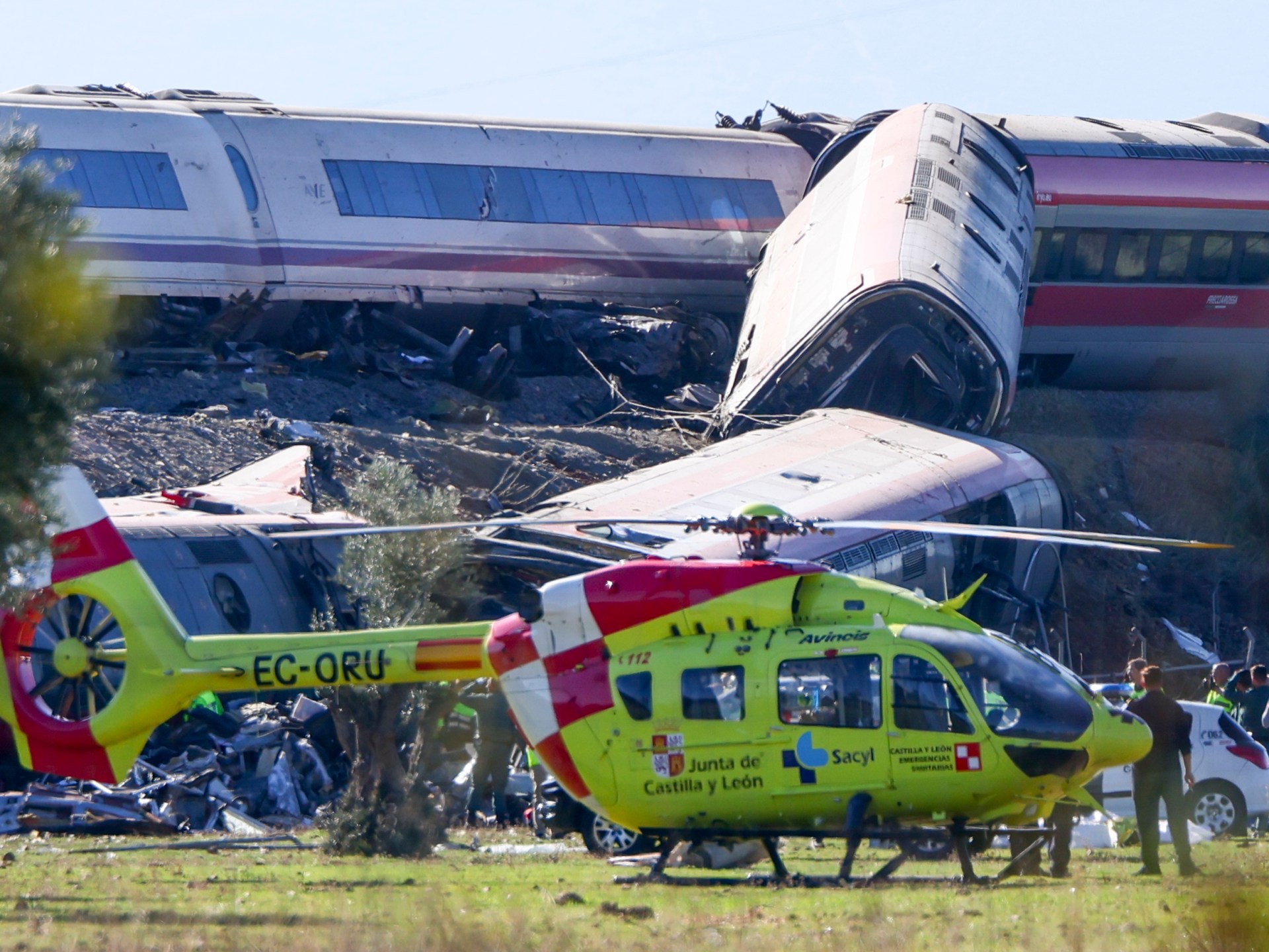 A yellow emergency helicopter is parked in front of derailed train carriages.